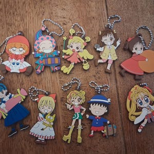 Könnte beinhalten: Eine Sammlung bunter Schlüsselanhänger mit Cartoonfiguren. Die Schlüsselanhänger zeigen verschiedene Charaktere in unterschiedlichen Outfits und Posen, mit silbernen Kettenbefestigungen. Die Charaktere haben große Augen und sind in leuchtenden Farben gehalten.