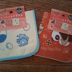 May include: Two square hand towels, each approximately 20 x 20 cm, featuring cartoon characters. One is light yellow with blue trim, the other is orange. Each towel has a tag with Japanese text and illustrations.