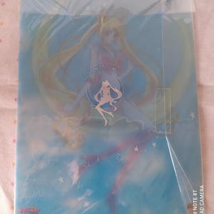K&ouml;nnte beinhalten: Ein durchsichtiger Plastikordner mit einer Sailor Moon-Illustration. Das Bild zeigt Sailor Moon in ihrer ikonischen Pose mit ihren langen blonden Haaren, die hinter ihr wehen. Der Hintergrund ist hellblau mit wei&szlig;en Sternen. Der Text "Sailor Moon" ist in einer stilisierten Schrift auf dem Ordner geschrieben.