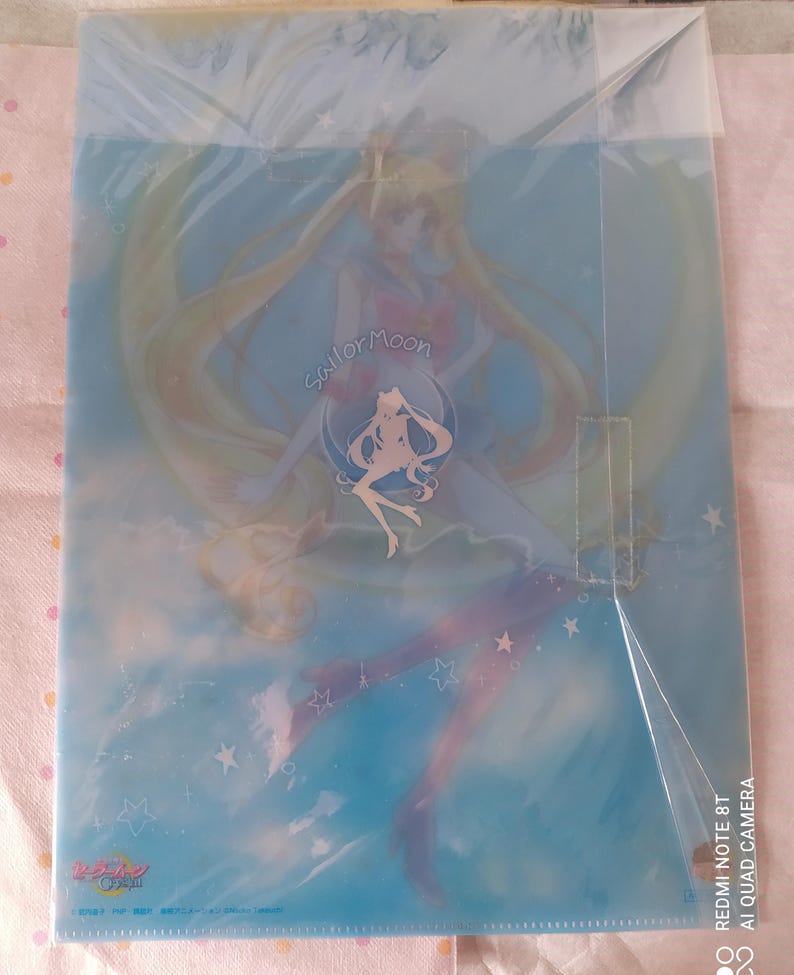 K&ouml;nnte beinhalten: Ein durchsichtiger Plastikordner mit einer Sailor Moon-Illustration. Das Bild zeigt Sailor Moon in ihrer ikonischen Pose mit ihren langen blonden Haaren, die hinter ihr wehen. Der Hintergrund ist hellblau mit wei&szlig;en Sternen. Der Text "Sailor Moon" ist in einer stilisierten Schrift auf dem Ordner geschrieben.