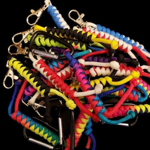 Può includere: Una collezione di portachiavi in paracord colorati con vari disegni e motivi. I portachiavi sono realizzati con diversi colori di paracord, tra cui rosso, blu, giallo, verde, bianco e nero. I portachiavi sono tutti attaccati a moschettoni argentati.