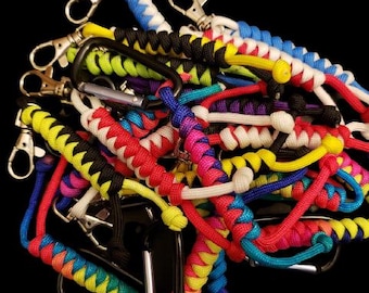 May include: A collection of colorful paracord keychains with various designs and patterns. The keychains are made with different colors of paracord, including red, blue, yellow, green, white, and black. The keychains are all attached to silver carabiners.