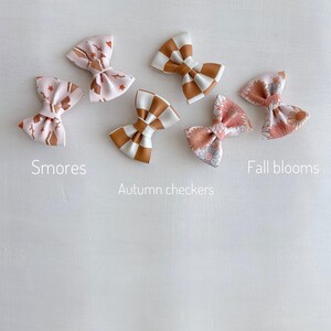 May include: Six fabric hair bows in various patterns and colours. The bows include designs labelled "Smores", "Autumn checkers", and "Fall blooms". The bows are in shades of pink, brown, white, and orange. The bows are arranged on a white surface.