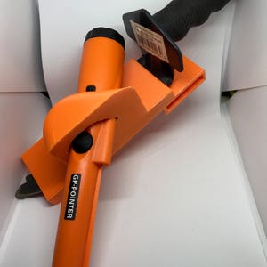 May include: An orange metal detector with a black button and the text "GP-POINTER" on the side. The detector is attached to a black and orange plastic holster with a black handle.