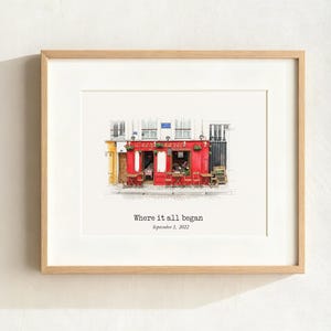 May include: Framed print of a red cafe with outdoor seating. The artwork features the text "Where it all began" and the date September 1, 2022. The frame is a light wood color.