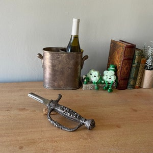 May include: An antique silver-toned bottle opener with an ornate handle, shaped like a sword, rests on a wooden surface. A wine bottle sits in a metal wine chiller. Decorative items include books and figurines.