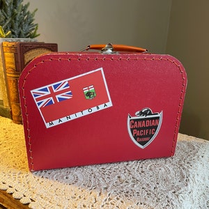 May include: Red vintage suitcase with a Canadian Pacific Railway logo, a Manitoba flag sticker, and a Canadian flag sticker.