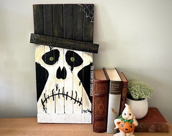 Puede incluir: Un letrero de madera blanco con una calavera pintada en blanco y negro que lleva un sombrero de copa negro. El letrero tiene el texto "The Country Cowshed" escrito verticalmente en el lado derecho. El letrero se apoya en dos libros y una pequeña figura de fantasma blanco con una calabaza.