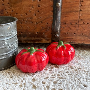 May include: Two glossy red ceramic tomato-shaped salt and pepper shakers with green stems, positioned next to a weathered gray metal measuring cup with embossed markings for 1/4 cup, 1/2 cup, 3/4 cup, and 1 cup measurements.