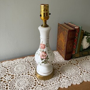 May include: White ceramic table lamp with a floral design featuring pink roses and green leaves. The lamp has a brass base and a gold-colored lamp holder. The lamp is on a wooden surface with a white lace runner.