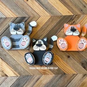 May include: Three wooden cat figurines in gray, black, and orange, each with a unique expression and paw details. The gray cat has striped markings, the black cat has a sad face, and the orange cat has a cheerful expression. The text on the image says "PLEASE NOTE LISTING IS FOR 1 CAT".