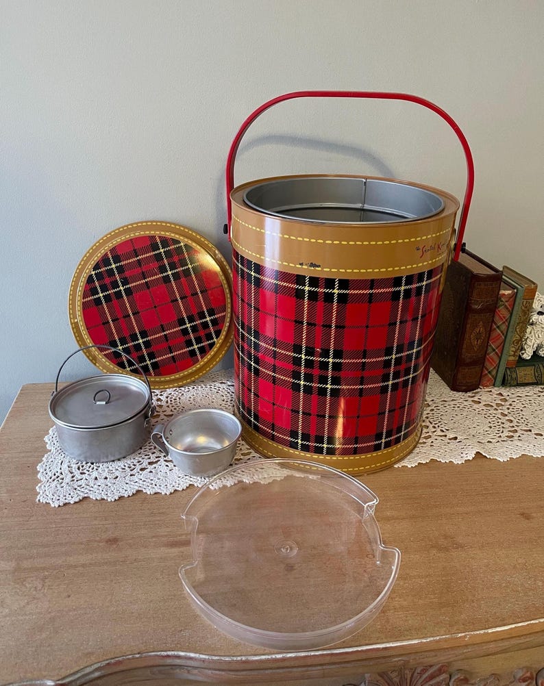Peut inclure: Une glaci&egrave;re de pique-nique vintage en m&eacute;tal rouge et noir &agrave; carreaux avec une poign&eacute;e rouge et un couvercle assorti. La glaci&egrave;re est sur une table en bois avec un plateau en plastique transparent, deux petites casseroles en aluminium avec couvercles et une dentelle blanche.