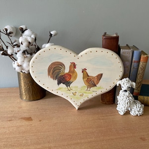 May include: A white heart-shaped wooden plaque with a painted scene of a rooster and hen with chicks. The plaque is surrounded by books and a cotton flower arrangement.