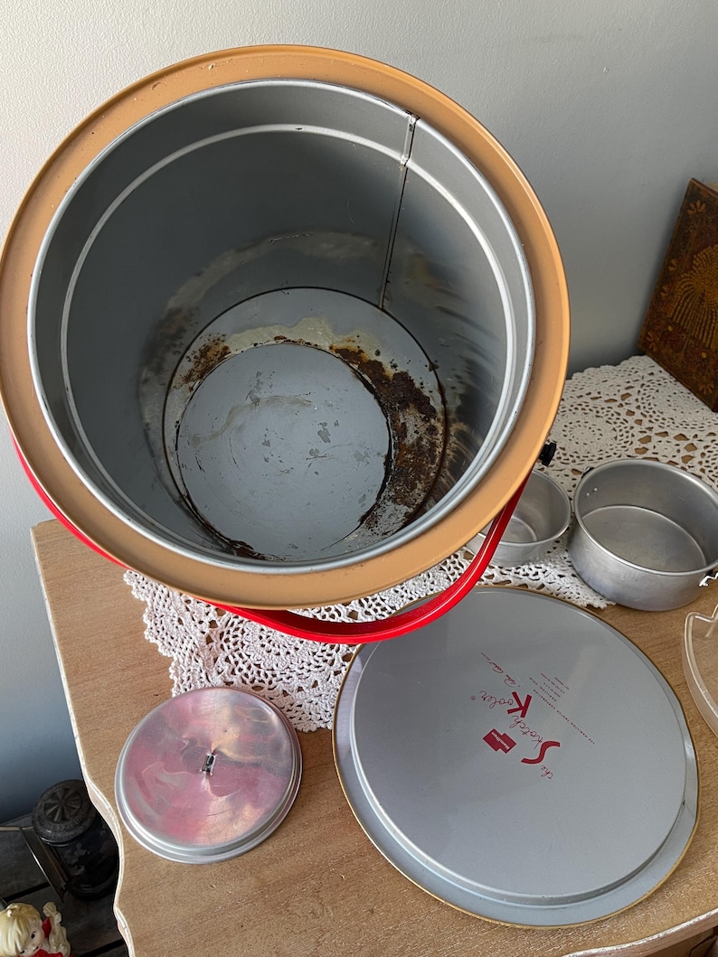 Peut inclure: Une glaci&egrave;re vintage en m&eacute;tal rouge et jaune avec un couvercle blanc. Le couvercle porte l'inscription "The Scotch Boy". La glaci&egrave;re est ouverte et vide.