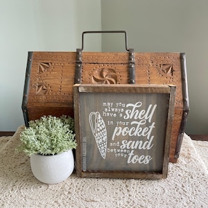 May include: A wooden sign with the text "may you always have a shell in your pocket and sand between your toes" and a seashell graphic. A wooden chest and a small potted plant are in the background.