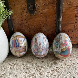 May include: Three decorative Easter egg-shaped tins with colorful illustrations. One tin features a flower market scene with a rabbit, another depicts rabbits in pastel clothing, and the third shows a rabbit with a basket. The tins are on a lace surface.