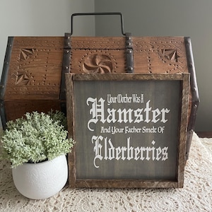 Puede incluir: Un letrero de madera enmarcado con el texto "Your Mother Was A Hamster And Your Father Smelt Of Elderberries" en letras blancas. El letrero está sobre un fondo gris. Un cofre de madera decorativo y una planta en maceta están en el fondo.