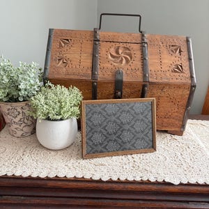 May include: A wooden trunk with metal accents and a handle, featuring carved geometric designs. A framed picture with a gray floral pattern sits in front of the trunk. Decorative plants in pots are on either side.