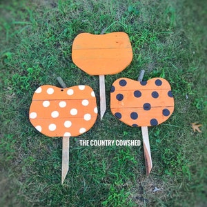 May include: Three wooden pumpkin signs with orange paint and black and white polka dots. The signs are on wooden stakes and are set in green grass. The text "THE COUNTRY COWSHED" is visible at the bottom of the image.