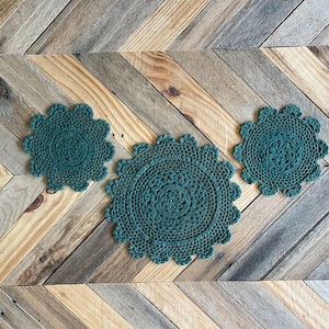 Puede incluir: Tres doilies de ganchillo en color verde oscuro, dispuestos sobre una superficie de madera con un patrón de chevron.