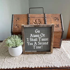 Op de afbeelding: Een rustiek houten bord met een zwarte achtergrond en witte tekst die "Go Away Or I Shall Taunt You A Second Time!" luidt. Het bord staat op een gehaakt tafellaken op een houten oppervlak. Het bord is gemaakt door The Country Cowshed.