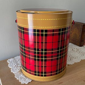 Peut inclure: Une glaci&egrave;re en m&eacute;tal rouge, noire et jaune &agrave; carreaux avec un couvercle brun et deux poign&eacute;es. La glaci&egrave;re est pos&eacute;e sur un napperon blanc en crochet.