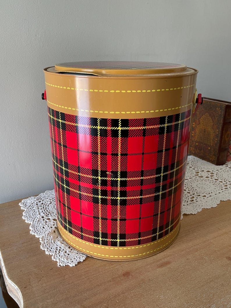 Peut inclure: Une glaci&egrave;re en m&eacute;tal rouge, noire et jaune &agrave; carreaux avec un couvercle brun et deux poign&eacute;es. La glaci&egrave;re est pos&eacute;e sur un napperon blanc en crochet.