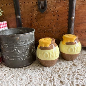 May include: A vintage-style kitchen scene featuring a metal measuring cup, two honey pot salt and pepper shakers, and a book. The salt and pepper shakers are yellow and brown with a honey design. The measuring cup has measurement markings.