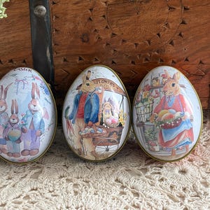 May include: Three decorative Easter egg-shaped tins with illustrations of rabbits. The tins have a gold trim and feature scenes of rabbits in an Easter setting. One tin has the word "Easter" on it.