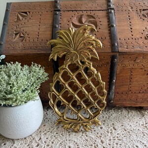 May include: A gold-coloured pineapple-shaped decorative object with a detailed leaf crown and a lattice design. The pineapple is resting on a cream-coloured crocheted surface, with a wooden chest in the background.