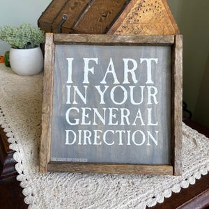 Op de afbeelding: Een rustiek houten bord met een grijze achtergrond en witte tekst die "I fart in your general direction" luidt. Het bord is ingelijst in een donkerhouten lijst en heeft de tekst "The Country Cowshed" in kleine witte letters onderaan.