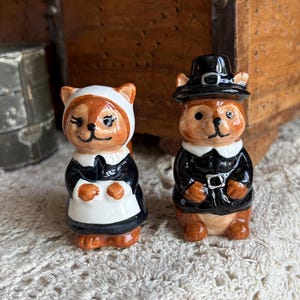 May include: Two ceramic squirrel figurines dressed in historical pilgrim attire. The left squirrel wears a white bonnet and black dress with a white apron. The right squirrel wears a black hat with a buckle and a black jacket with a white collar.