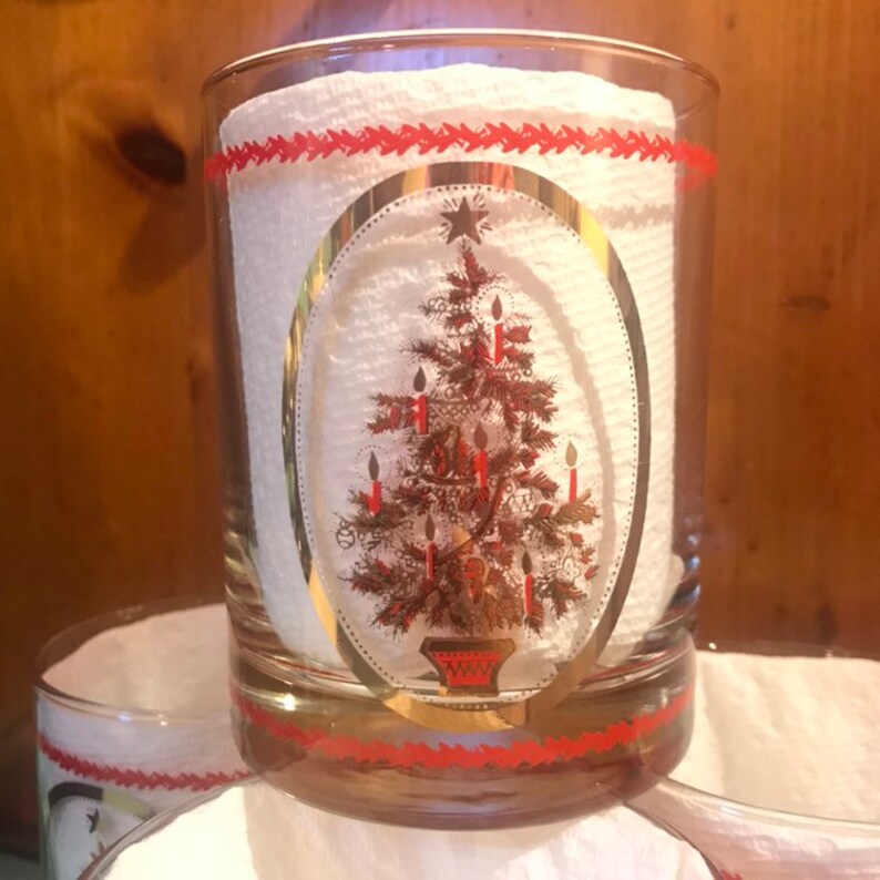 May include: Set of four clear glass tumblers with a gold rim and a red band. Each tumbler features a gold-outlined oval with a Christmas tree design. The tree is decorated with red candles and gold ornaments.