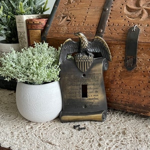 Könnte beinhalten: Ein goldfarbener Lichtschalter aus Metall mit Adler- und Schriftrollen-Design. Die Schriftrolle zeigt den Text "Freiheit ist das Recht zu wählen, das Recht, für sich selbst Alternativen der Wahl zu schaffen. ... ein Mensch nicht als Instrument, eine Sache." - Thomas Jefferson