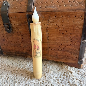 May include: A battery-operated Winnie the Pooh candle with a flickering LED flame. The candle has a cream-colored wax appearance with a printed image of Winnie the Pooh and the word "Hunny". The candle is placed in front of a wooden chest.