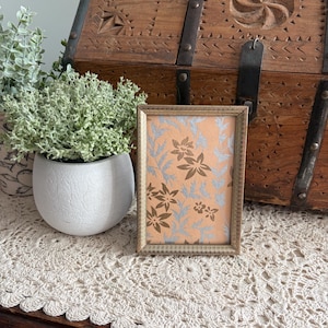 May include: A small, rectangular gold-colored picture frame with a floral design on peach-colored paper. The frame is placed on a cream-colored, crocheted doily. A white pot with greenery and a wooden chest are in the background.