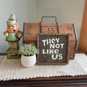 May include: A wooden sign with the text "They Not Like Us" in white lettering on a dark brown background. The sign is sitting on a table with a small potted plant and a leprechaun figurine.