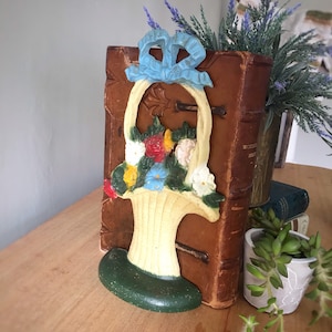 May include: A cast iron doorstop shaped like a basket of flowers. The basket is painted white with a green base and a blue bow. The flowers are red, yellow, white, and blue.