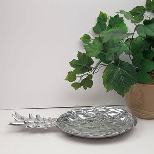 May include: A silver-colored pineapple-shaped serving dish. The dish has a textured surface and a detailed design, with a stem and leaves at one end. The dish is on a white surface, with a potted plant in the background.