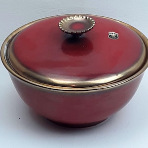 May include: A red ceramic serving bowl with a lid. The bowl has a gold rim and a gold handle on the lid. A small emblem is on the lid. The bowl is likely vintage and suitable for serving food.