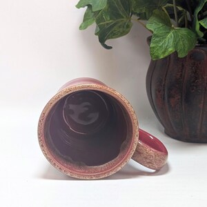 May include: A ceramic mug with a deep magenta interior and a textured, speckled beige and brown rim. The mug features a matching speckled handle extending from the side, viewed from a top-down perspective, showcasing the cylindrical shape.