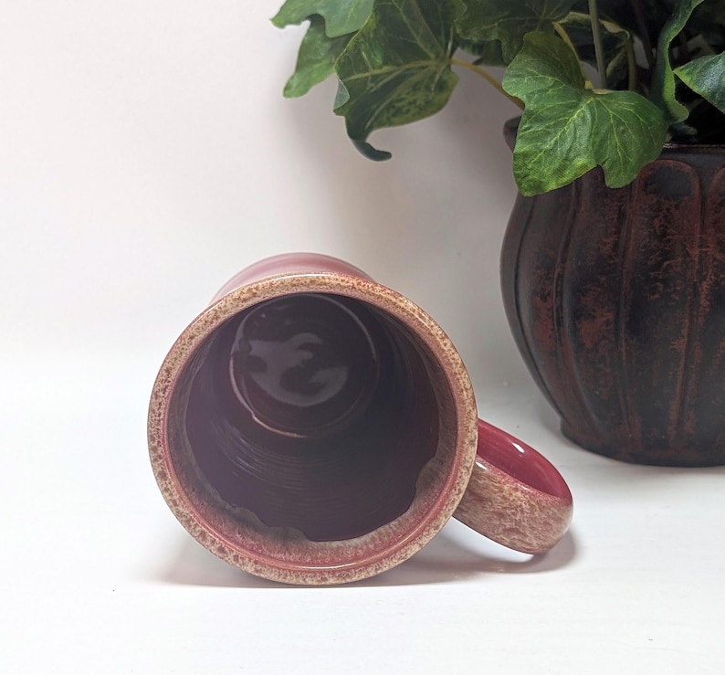 May include: A ceramic mug with a deep magenta interior and a textured, speckled beige and brown rim. The mug features a matching speckled handle extending from the side, viewed from a top-down perspective, showcasing the cylindrical shape.