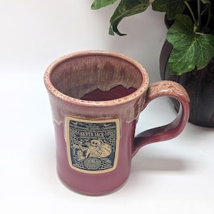 May include: A ceramic mug with a dusty rose glaze and a lighter, mottled rim. A rectangular label on the front features a black and white illustration of Santa Jack from The Nightmare Before Christmas, with text reading Santa Jack, Cranberry Creme Br&ucirc;l&eacute;e, and Bones.