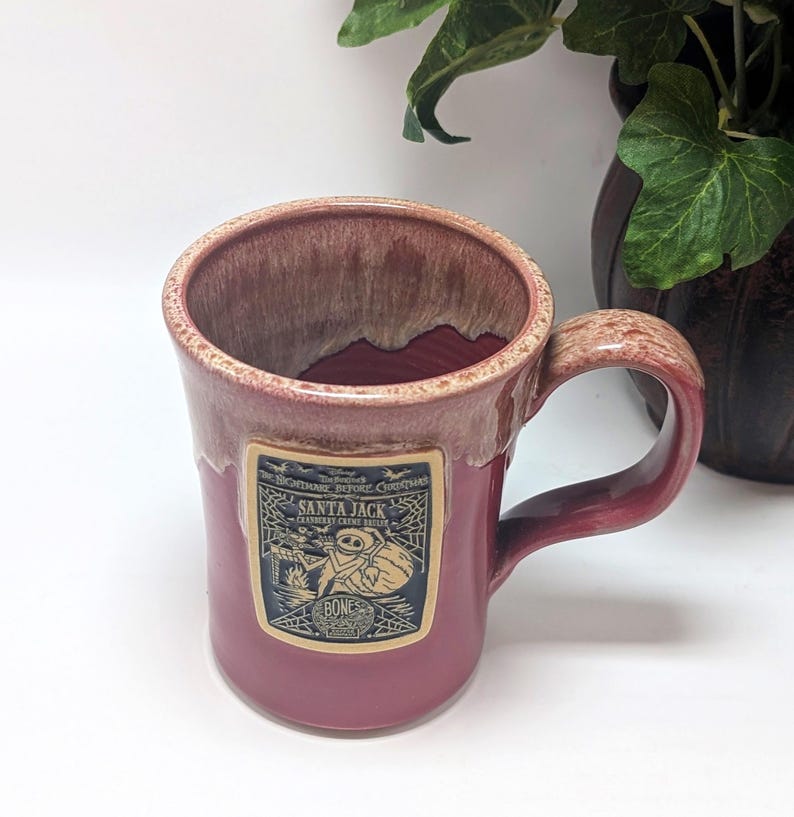 May include: A ceramic mug with a dusty rose glaze and a lighter, mottled rim. A rectangular label on the front features a black and white illustration of Santa Jack from The Nightmare Before Christmas, with text reading Santa Jack, Cranberry Creme Br&ucirc;l&eacute;e, and Bones.