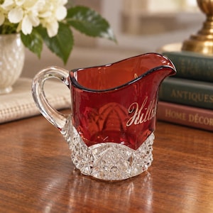May include: A small glass creamer pitcher featuring a deep red upper section with the script text Hillside and a clear, faceted, diamond-patterned base. The pitcher includes a curved clear glass handle and a distinct pouring spout.