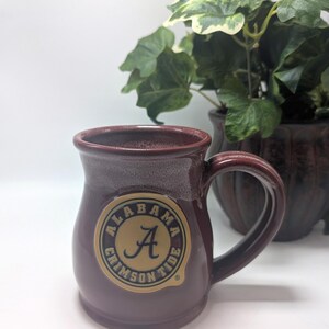 May include: A dark maroon ceramic mug with a rounded body and a curved handle. The front features a circular emblem with a gold background, a black border, and the text Alabama Crimson Tide surrounding a stylized black letter A in the center.