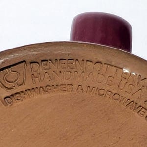 May include: The underside of a tan ceramic mug featuring a dark maroon handle. Embossed text reads DENEENPOTTERY.COM HANDMADE USA 2022 DISHWASHER & MICROWAVE SAFE, accompanied by a logo depicting a stylized vessel shape with a central circle.