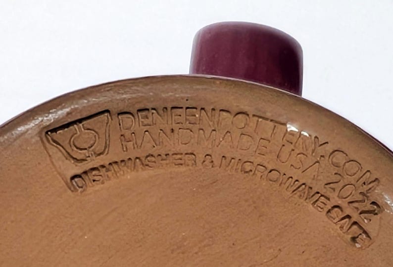 May include: The underside of a tan ceramic mug featuring a dark maroon handle. Embossed text reads DENEENPOTTERY.COM HANDMADE USA 2022 DISHWASHER & MICROWAVE SAFE, accompanied by a logo depicting a stylized vessel shape with a central circle.