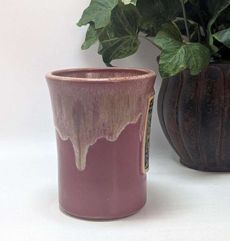 May include: A cylindrical ceramic mug featuring a solid mauve base and a lighter, cream-colored glaze that drips down from the rim. The mug has a smooth, glossy finish and a straight-sided profile, showcasing a distinct two-tone color transition.