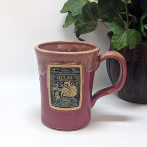May include: A ceramic mug with a glossy cranberry red glaze and a lighter drip effect at the rim. A rectangular label features a black and cream graphic of Santa Jack from The Nightmare Before Christmas, including text and a spiderweb design.